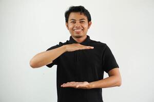 Jeune asiatique homme contre une blanc studio Contexte portant noir polo t chemise, en portant quelque chose avec tous les deux mains, produit présentation. photo