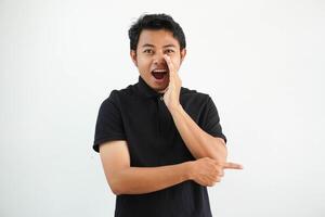 Jeune asiatique homme posant sur une blanc toile de fond en disant une potins, montrer du doigt à côté rapports quelque chose, portant noir polo t chemise. photo