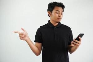 Jeune asiatique homme en portant une mobile téléphone isolé sur blanc Contexte sérieux et montrer du doigt de côté, montrant quelque chose à Vide espace, portant noir polo t chemise. photo