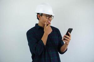 Jeune asiatique Masculin architecte ingénieur souriant en portant le sien menton en pensant tandis que à la recherche à le téléphone portable il a été en portant isolé sur blanc Contexte. photo