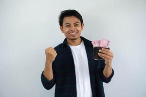 Jeune asiatique homme montrant excité visage expression tandis que en portant le sien portefeuille plein de argent avec serré les poings isolé sur blanc Contexte photo