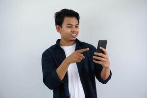 souriant Jeune asiatique homme lorsque à la recherche à le sien mobile téléphone et point avec une doigt isolé sur blanc Contexte photo