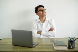 Jeune asiatique homme d'affaire dans une lieu de travail à la recherche en haut tandis que souriant, portant blanc chemise avec des lunettes isolé photo