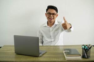 Jeune asiatique homme d'affaire dans une lieu de travail avec les pouces en haut car quelque chose bien a arrivé, portant blanc chemise avec des lunettes isolé photo