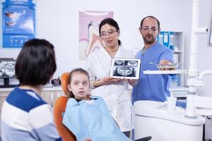 dentiste discuter à propos cavité la prévention avec peu fille parent stomatologue expliquant les dents diagnostic à mère de enfant dans santé clinique en portant radiographie. photo