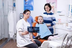 dentisterie spécialiste parlant à propos cavité procédure à enfant porter dentaire bavoir. stomatologue expliquant les dents diagnostic à mère de enfant dans santé clinique en portant radiographie. photo