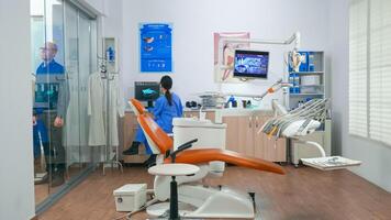 infirmière attrayant suivant patient dans stomatologie pièce montrant à mensonge sur chaise. dentisterie assistant séance dans consultation dentaire pièce avec personnes âgées femme tandis que médecin Parlant avec vieux homme dans Contexte. photo