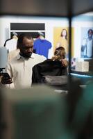 africain américain homme achats pour chemise et vérification vêtements dans Stock en utilisant téléphone intelligent en ligne application. Vêtements boutique client en portant tenue sur grille et défilement site Internet sur mobile téléphone photo