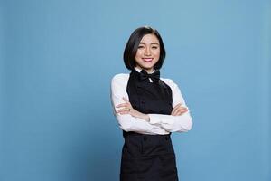 souriant Jeune attrayant serveuse habillé dans restaurant uniforme permanent avec bras franchi portrait. de bonne humeur femme réceptionniste posant avec plié mains et à la recherche à caméra photo