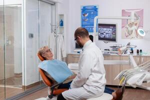 dentiste dans dentaire clinique en disant diagnostique à Sénior femme portant laboratoire manteau. médical les dents se soucier preneur parlant avec patient séance sur chaise pendant consultation. photo