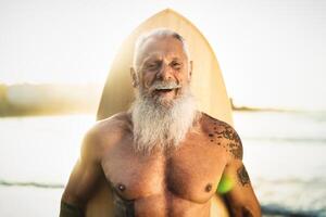 en forme Sénior Masculin ayant amusement surfant pendant le coucher du soleil temps - retraité homme formation avec planche de surf sur le plage - personnes âgées en bonne santé gens mode de vie et extrême sport concept photo