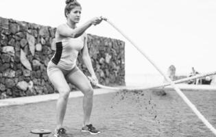 en forme femme formation avec bataille corde Extérieur - femelle athlète Faire bras des exercices sur le plage - corps bâtiment et sport mode de vie concept - noir et blanc Éditer photo