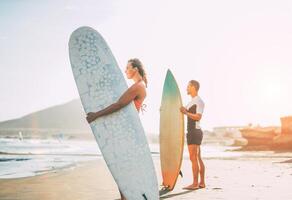 Jeune couple de surfeurs permanent sur le plage avec planches de surf en train de préparer à le surf sur haute vagues pendant une magnifique le coucher du soleil - personnes, mode de vie, sport concept - concentrer sur femme photo