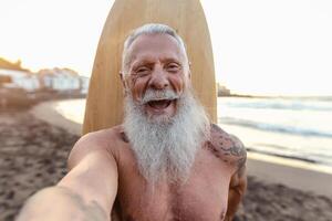 content Sénior surfeur prise selfie tandis que ayant amusement surfant à le coucher du soleil temps - personnes âgées santé gens mode de vie et extrême sport concept photo