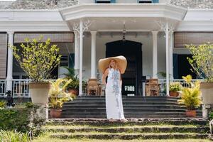 le femme dans le gros chapeau est souriant. une magnifique fille dans une gros chapeau et blanc robe sourit à l'extérieur un vieux colonial bâtiment sur le île de maurice photo