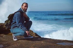 barbu Masculin voyageur aventurier avec sac à dos, séance sur le bord de une rocheux falaise contre rupture vagues Contexte photo