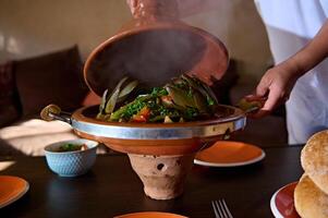 fermer mains de une femme au foyer ouverture le argile couvercle de marocain tajine tandis que portion table pour dîner à Accueil photo