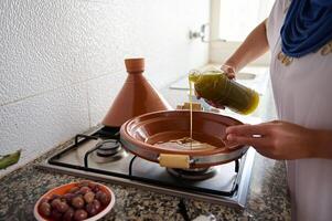 fermer femme au foyer début cuisine repas dans tajine argile pot, verser supplémentaire vierge olive pétrole à l'intérieur une argile plat photo