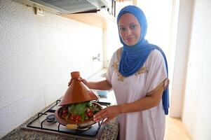 souriant milieu est femme au foyer avec tête couvert dans hijab, permanent par poêle, cuisine marocain tajine avec légumes photo