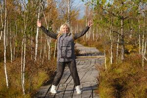 une content femme avec une sac à dos sauts sur une en bois chemin dans une marais dans Elnya, biélorussie photo