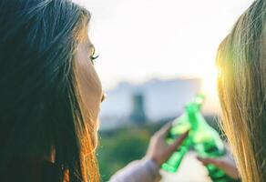 Jeune femmes grillage Bière sur une le coucher du soleil - content copains en buvant et applaudissement bières dans une parc Extérieur admiratif le magnifique vue - personnes, boissons, relation amicale concept - concentrer sur femelle visage sur le la gauche photo