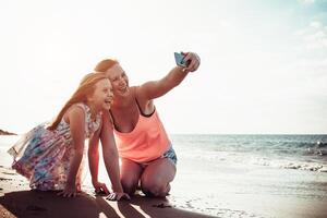 mère et fille prise selfie avec mobile téléphone intelligent tandis que en jouant sur tropical plage à le coucher du soleil - content famille ayant amusement avec Nouveau La technologie applications pour social médias - parentalité concept photo