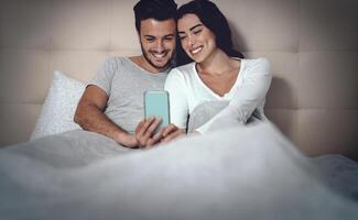 Jeune couple en utilisant mobile téléphone dans lit - homme et femme en train de regarder téléphone intelligent et ayant amusement avec social réseaux La technologie applications - jeunesse millénaire et gens relation concept photo