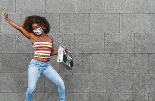 Jeune afro femme portant visage masque dansant Extérieur tandis que écoute à la musique avec sans fil écouteurs et ancien boombox stéréo photo