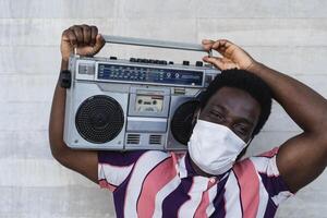 Jeune africain homme en portant ancien boombox ayant amusement Extérieur - noir homme portant visage masque tandis que écoute la musique avec stéréo pendant couronne virus pandémie - jeunesse gens et santé se soucier concept photo