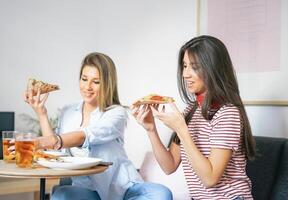 Jeune femmes en mangeant vite nourriture Pizza et en buvant Bière à Accueil - content copains profiter une dîner séance sur canapé dans le vivant pièce dans leur appartement - gens à manger concept photo