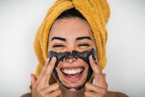 Jeune souriant femme appliquant charbon masque sur visage - content fille ayant peau se soucier spa journée à Accueil - en bonne santé beauté nettoyer traitement et jeunesse gens mode de vie concept photo