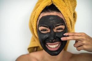 Jeune souriant femme appliquant charbon masque sur visage - content fille ayant peau se soucier spa journée à Accueil - en bonne santé beauté nettoyer faciale traitement et jeunesse gens mode de vie concept photo