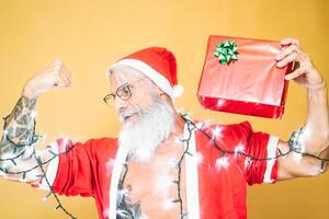 content tatoué Père Noël claus équipé avec blanc lumières donnant Noël cadeau - branché barbe Sénior portant Noël vêtements et en portant présent - personnes, amusement et vacances concept photo