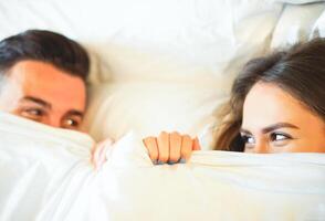 Jeune espiègle couple ayant amusement dans le lit - content les amoureux à la recherche timide à chaque autre dans yeux mensonge en dessous de blanc feuilles dans te chambre à Accueil - marrant et romantique aimant concepts - concentrer sur femme œil photo