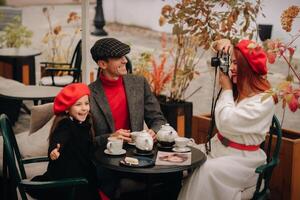 une élégant famille de Trois est séance à une table à l'extérieur dans une café et en buvant café. papa, maman et fille dans le l'automne ville photo