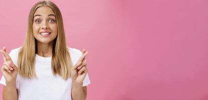 optimiste optimiste excité fille avec bronzé peau et tout droit juste cheveux looing excité et content à caméra souriant avec espérer traversée les doigts pour bien la chance fabrication souhait posant contre rose mur photo