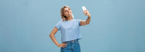 élégant mignonne femelle Gym directeur prise selfie avec Nouveau téléphone intelligent posant avec vaste heureux sourire et en portant main sur hanche fabrication Publier pour suiveurs permanent insouciant et détendu plus de bleu mur photo