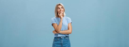 rêveur réfléchi et Créatif artistique blond femme dans denim short et T-shirt souriant avec curiosité émouvant lèvre et regarder à plus haut la gauche coin sentiment nostalgique, en pensant plus de bleu mur photo