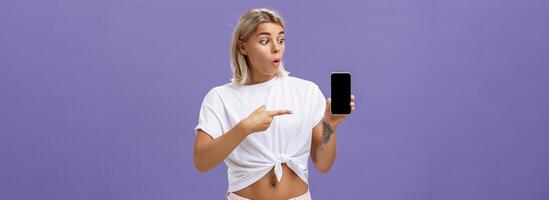 à la taille coup de impressionné et Enchanté magnifique européen femelle avec blond cheveux et tatouages à la recherche et montrer du doigt à impressionnant marque Nouveau téléphone intelligent permanent plus de violet Contexte photo