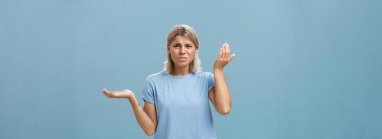 wtf vous en disant. portrait de énervé et confus élégant petite amie avec blond cheveux élevage paume haute et de côté avec perplexe expression étant interrogé audition stupide absurdité plus de bleu mur photo