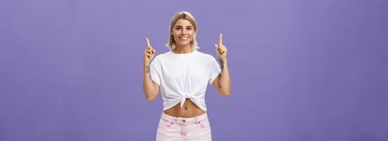 temps Regardez vers le haut et bouge toi vers l'avant entreprise échelle. portrait de attrayant ambitieux et élégant Jeune blond femme avec tatouages et percé ventre montrer du doigt en haut et souriant largement plus de violet mur photo