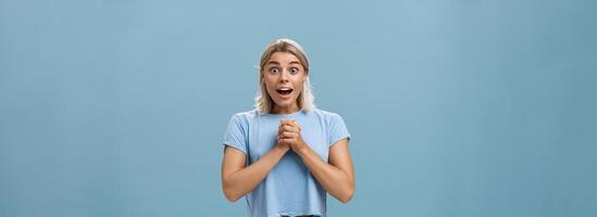 vraiment est il pour moi. portrait de surpris reconnaissant attrayant femme avec blond la Coupe de cheveux serrant mains ensemble sur Sein ouverture bouche halètement de admiration et fascination permanent plus de bleu mur photo