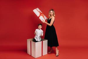 maman ouvre une énorme Noël cadeau dans lequel une garçon des stands et spectacles le classe sur une rouge Contexte photo