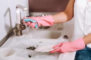 femme au foyer fille dans rose gants lavages vaisselle par main dans le évier avec détergent. le fille nettoie le maison et lavages vaisselle dans gants à maison. photo