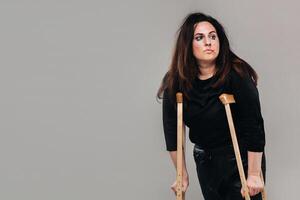 une femme battu dans noir vêtements avec béquilles dans sa mains sur une gris Contexte photo