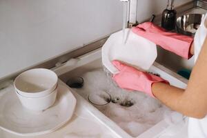 femme au foyer fille dans rose gants lavages vaisselle par main dans le évier avec détergent. le fille nettoie le maison et lavages vaisselle dans gants à maison. photo