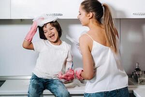 femme au foyer maman dans rose gants lavages vaisselle avec sa fils par main dans le évier avec détergent. une fille dans blanc et une enfant avec une jeter nettoie le maison et lavages vaisselle dans fait maison rose gants. photo