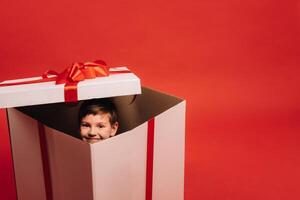 une peu garçon est assis dans une Noël présent et regards en dehors de il sur une rouge Contexte photo