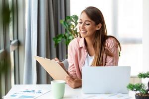 travail à distance, technologie et concept de personnes - jeune femme d'affaires souriante et heureuse avec ordinateur portable et papiers travaillant au bureau à domicile photo