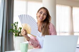 fatigué surchauffé Jeune femme tenir vague ventilateur souffrir de chaleur transpiration intérieur travail sur portable à Accueil bureau, agacé femme ressentir inconfortable chaud été temps problème non air Conditionneur concept photo